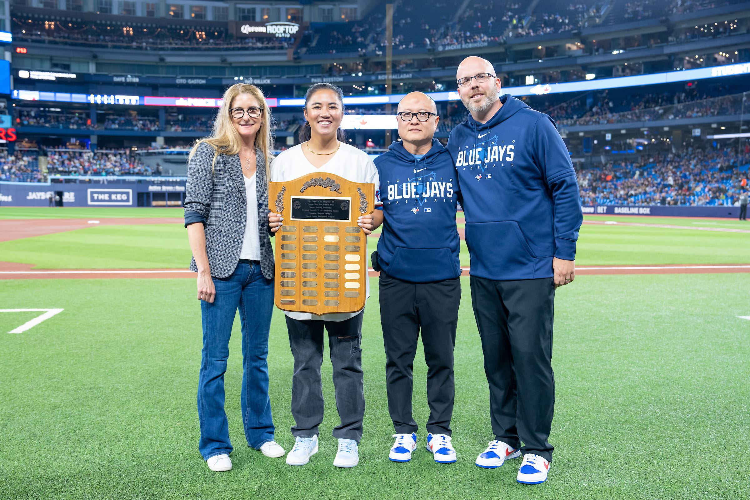 Sheridan athletic therapy graduate Katie Reyes stands on the Rogers Centre field with a plaque she received for serving as the Toronto Blue Jays' athletic therapy intern during the 2022 season. Reyes was joined on the field by Sheridan athletic therapy field placement coordinator Julie Dickson and Blue Jays assistant trainers Voon Chong and Drew MacDonald.
